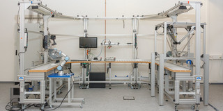 Forschungsdemonstrator des Projekts ViKIMon: hybrides Montagesystem mit KI-gestützter Qualitätsprüfung U-förmige Montagelinie aus Aluminiumprofilen in der IETC-Laborfläche. Links steht ein Roboterarm, rechts ein Deltaroboter. In der Mitte befinden sich ein Bildschirm, Werkstückträger auf einem Transportband und oberhalb davon eine Prüfstation.