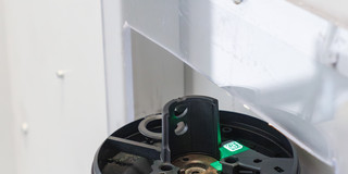Nahaufnahme eines technischen Prüfgeräts in einem Labor: Ein rundes schwarzes Bauteil liegt eingespannt auf einer Metallplatte, daneben ist ein kleiner grüner Sensor mit Licht zu sehen